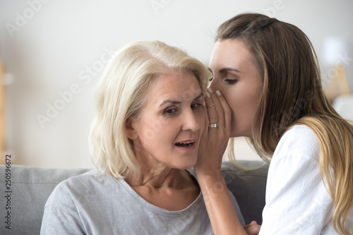 Woman sharing secret with surprised aged mother, whispering in ear