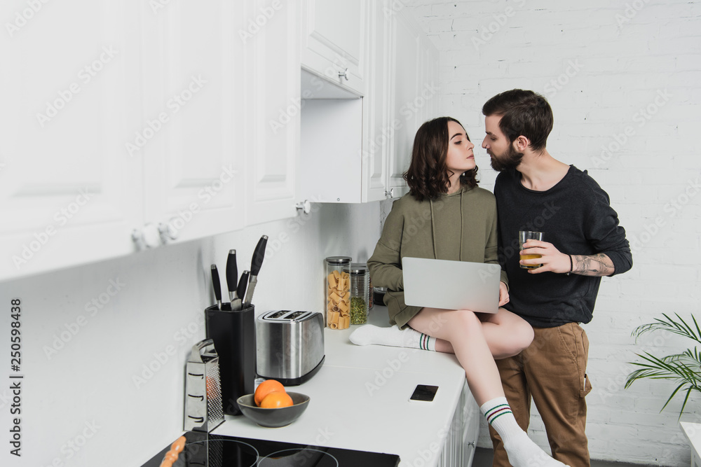 beautiful couple with orange juice and laptop looking at each other during breakfast in kitchen beautiful couple with orange juice and laptop looking at each other during breakfast in kitchen