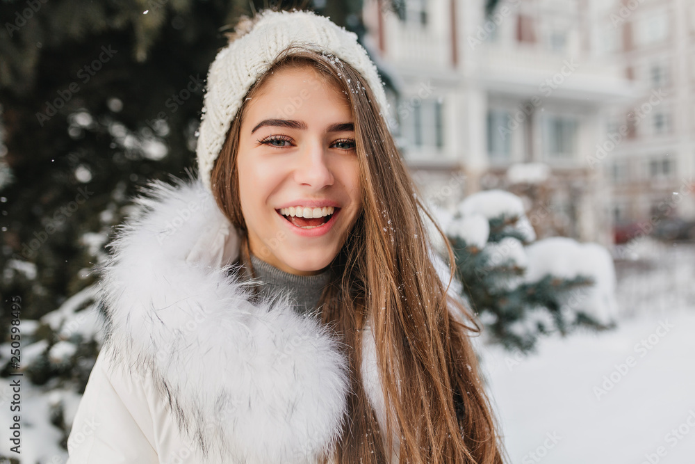 Closeup portrait of joyful smiling girl in knitted hat posing outdoor on street full of snow. Cheerful blonde lady with blue eyes enjoying winter time spending weekend in the yard.. Closeup portrait of joyful smiling girl in knitted hat posing outdoor on street full of snow. Cheerful blonde lady with blue eyes enjoying winter time spending weekend in the yard..