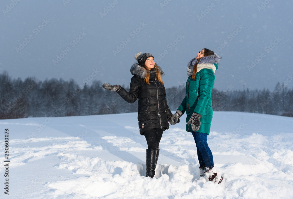 Two happy Caucasian girls are holding hands and looking at the evening sky during snowfall in rural.