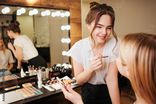 Photos Cheerful young make up artist stand in front of blonde woman in beauty room and smile