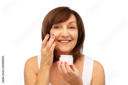 beauty, skincare and cosmetics concept - smiling senior woman applying cream to her face