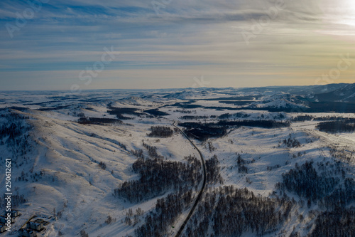 Aerial; drone panoramic view of winter mountain landscape with blue sky and clouds; sunny day in russian countryside; snow fields and mixed forest with birches and pine trees; local ski resort Bannoe