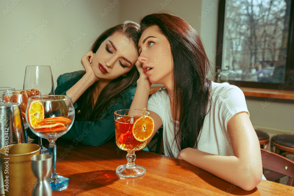 Female sad and tired friends having a drinks at bar. They are sitting at a wooden table with cocktails. They are wearing casual clothes. Female sad and tired friends having a drinks at bar. They are sitting at a wooden table with cocktails. They are wearing casual clothes.