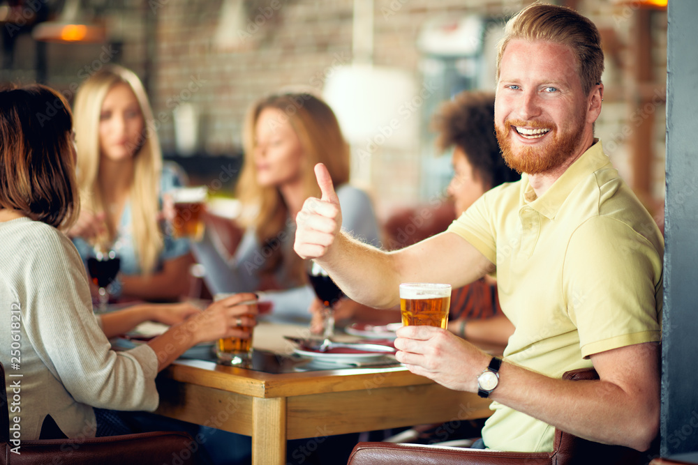 Man drinking beer and giving thumbs up while sitting in restaurant. In background his friends chatting and drinking alcohol.