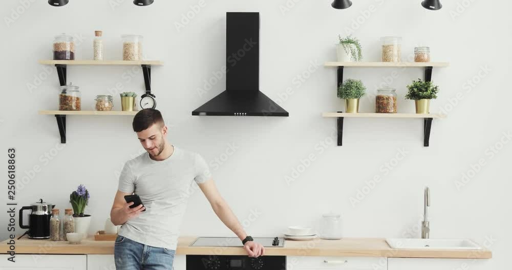 Smart phone man browsing on mobile phone at home kitchen. Handsome young man browsing on smartphone smiling happy. Smart phone man browsing on mobile phone at home kitchen. Handsome young man browsing on smartphone smiling happy.