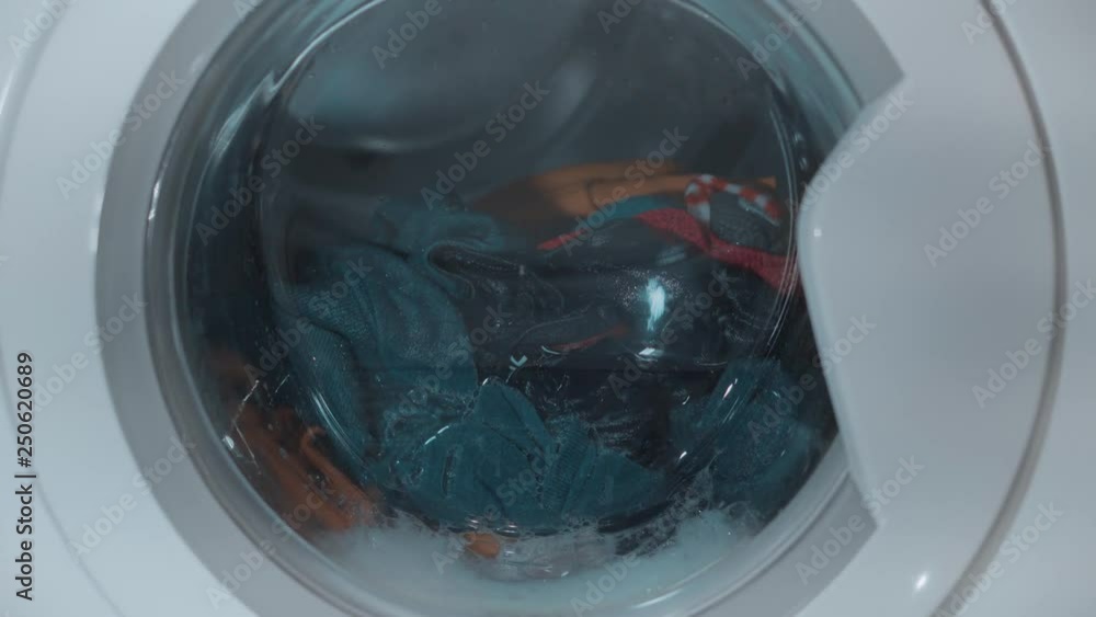 Womam is gently putting colourful laundry into washing machine. A young girl closes the door of the washing machine foot. The washing machine drum washes clothes. 4k video. Womam is gently putting colourful laundry into washing machine. A young girl closes the door of the washing machine foot. The washing machine drum washes clothes. 4k video.