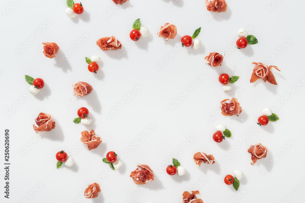 flat lay of tasty prosciutto near red cherry tomatoes, mozzarella cheese and green basil leaves on white background flat lay of tasty prosciutto near red cherry tomatoes, mozzarella cheese and green basil leaves on white background