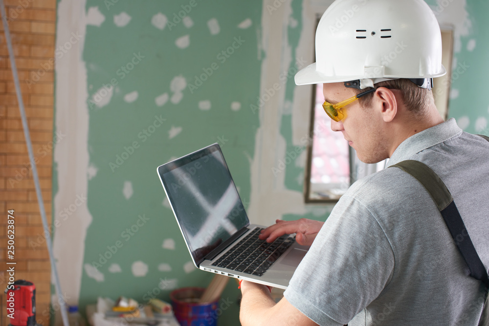 Room repair. Builder in helmet and glasses stands with a laptop in his hands on the background of the construction Room repair. Builder in helmet and glasses stands with a laptop in his hands on the background of the construction