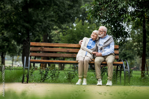 smiling senior couple embracing and holding hands while resting on wooden bench in park