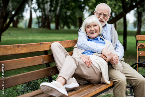 happy senior woman laying on wooden bunch near smiling husband in park