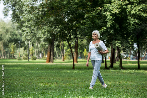 smiling senior woman holding fitness mat while walking in park