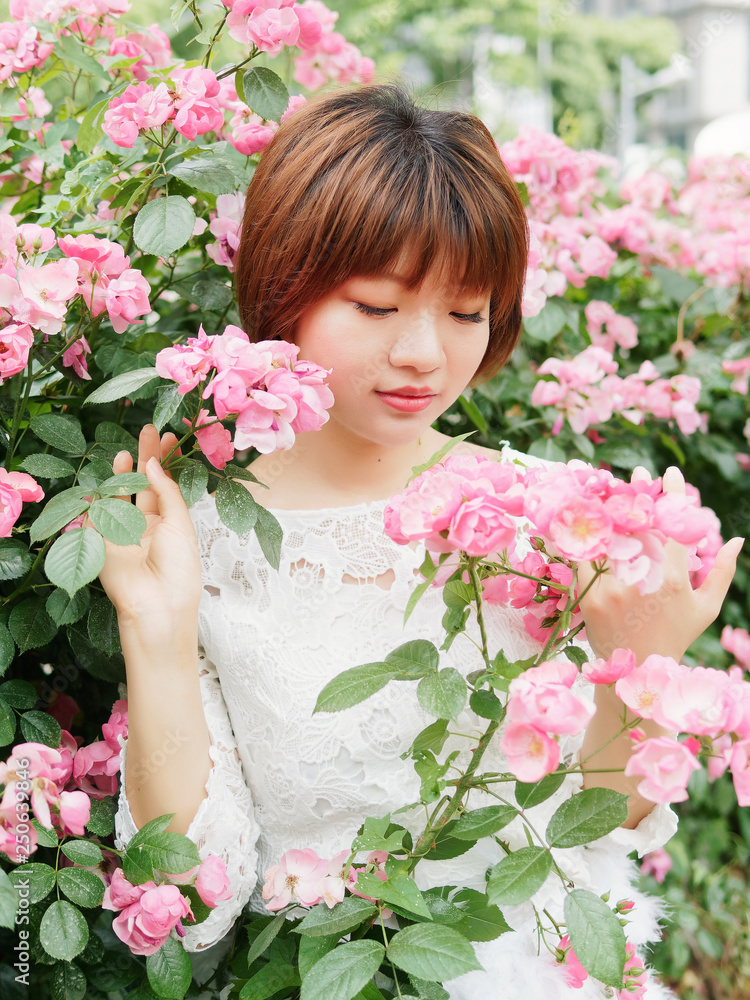 Fototapeta premium Beautiful young brunette woman in white lace dress looking and smiling in rose garden. Outdoor fashion portrait of glamour Chinese stylish girl. Emotions, people, beauty and lifestyle concept.