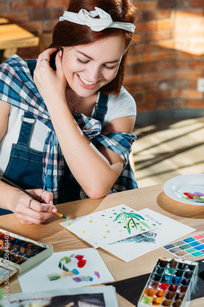 Fototapeta premium Studio workplace. Painting hobby. Smiling redhead woman doing first brushstrokes with palette supplies around.