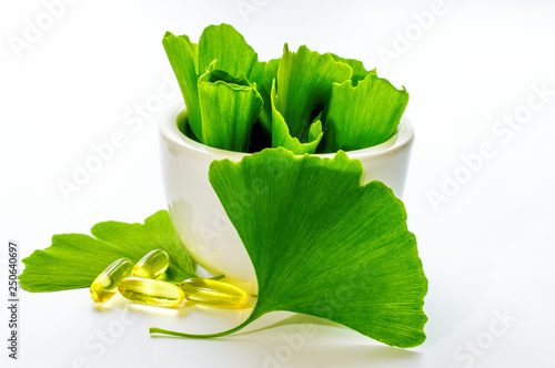 Healing leaves of ginkgo biloba tree in a white ceramic bowl and yellow capsules. Green leaves on a white background.	