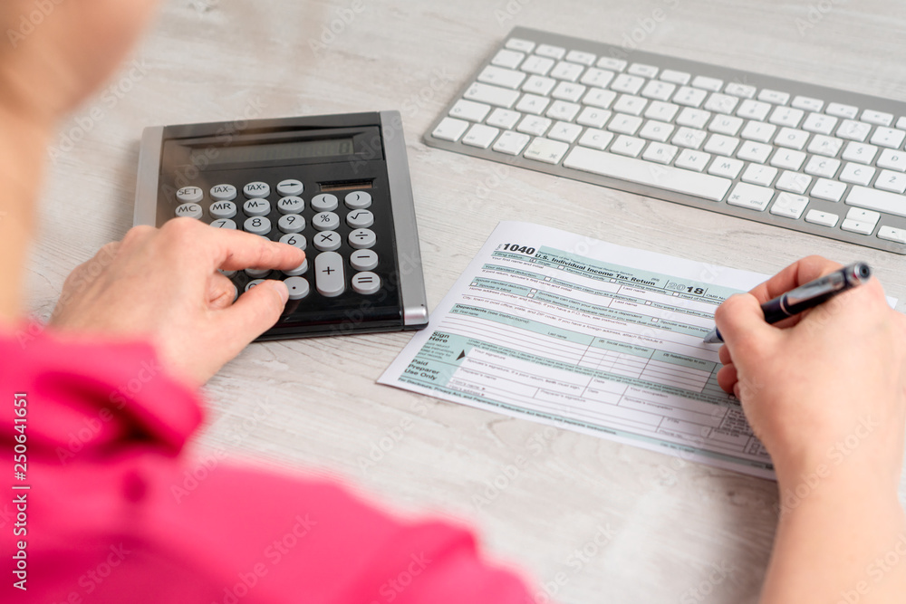 Over the shoulder photo of woman calculating and filling US tax form next to computer keyboard and tax form 1040. 15th April 2019 tax day Over the shoulder photo of woman calculating and filling US tax form next to computer keyboard and tax form 1040. 15th April 2019 tax day