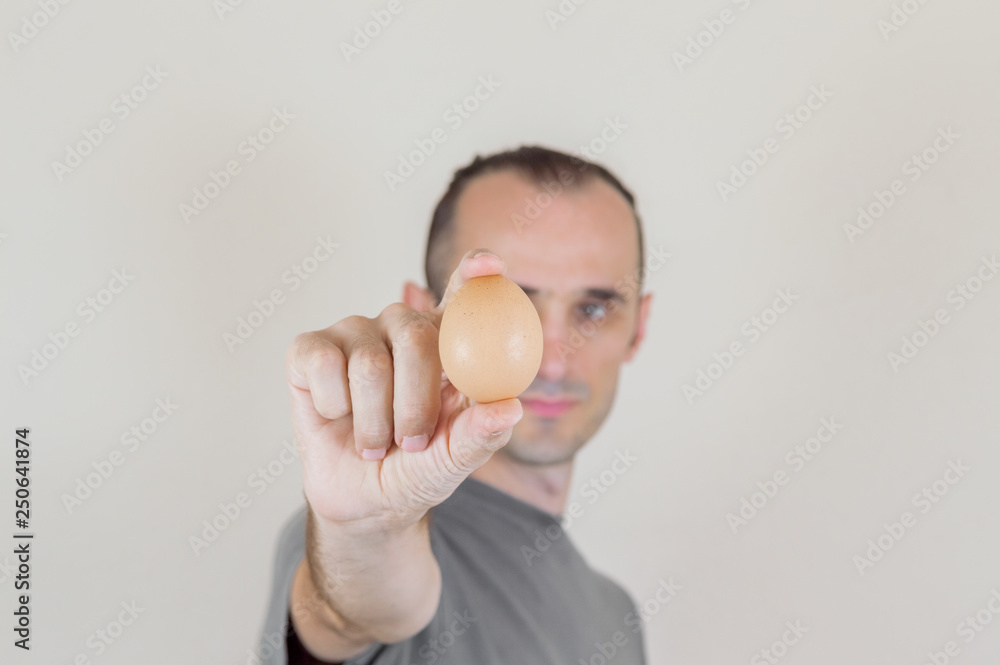Caucasian man with gray tshirt holding and showing an egg between his fingers, blurry face on white background Caucasian man with gray tshirt holding and showing an egg between his fingers, blurry face on white background