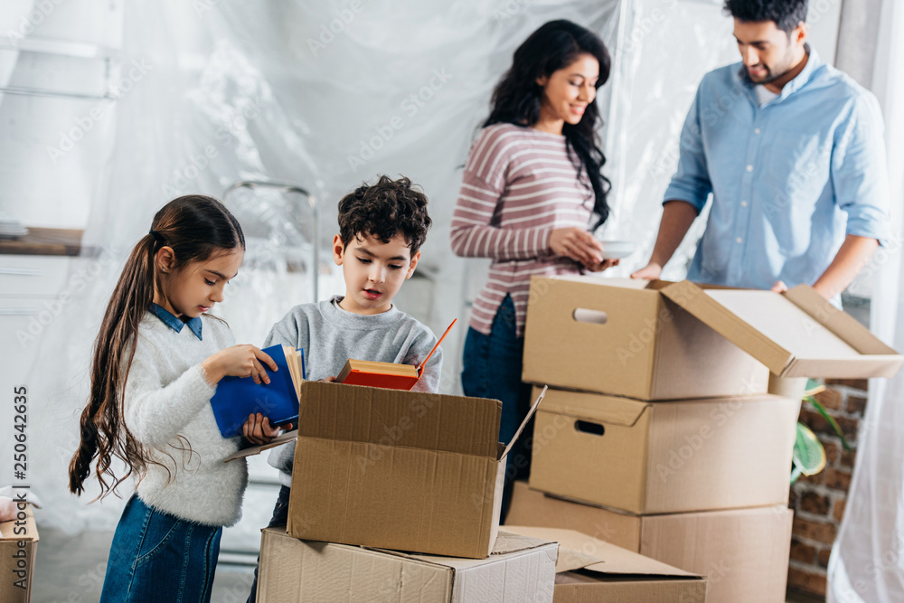 selective focus of cute kids holding books while parents standing near boxes in new home selective focus of cute kids holding books while parents standing near boxes in new home