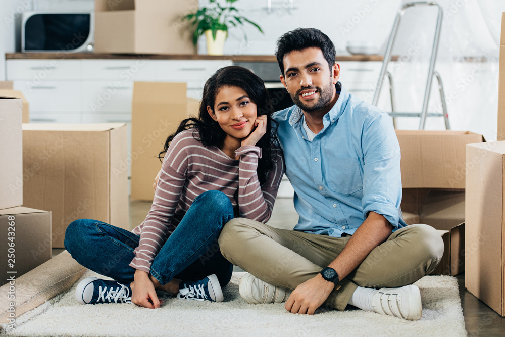 cheerful latin couple sitting with crossed legs near boxes in new home cheerful latin couple sitting with crossed legs near boxes in new home