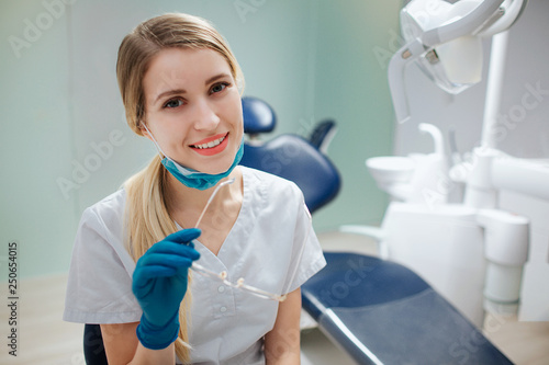 Cheerful young female dentist sit in room and pose to camera. She smile. Doctor hold glasses in hand.