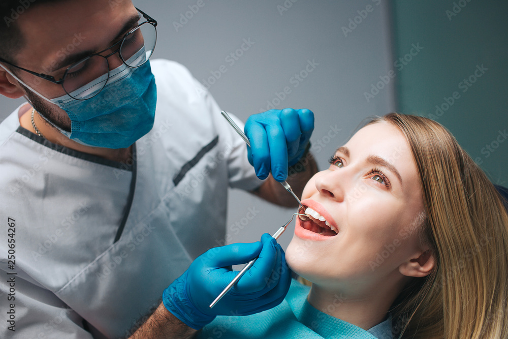 Close up of dentist checking client's teeth with special tools. Young woman sit in chair in room and keep mouth opened. She look up. Close up of dentist checking client's teeth with special tools. Young woman sit in chair in room and keep mouth opened. She look up.