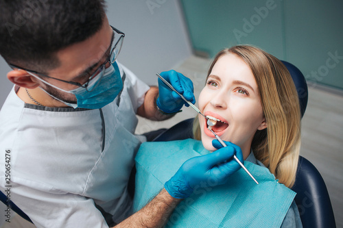 Serious dentist in mask and white robe stand beside client and checking teeth condition. Young woman sit in chair and keep mouth opened. She look up.