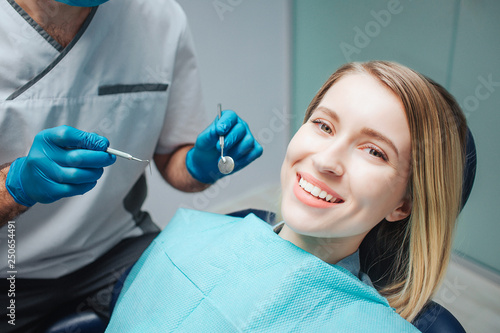 Positive young woman sit in chairin dentistry and smile to camera. She show beautiful smile. Doctor hold tools for teeth treatment.