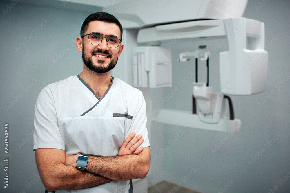 Nice cheerful dentist stand in x-ray room and pose on camera. He smile. X-ray machine behind.