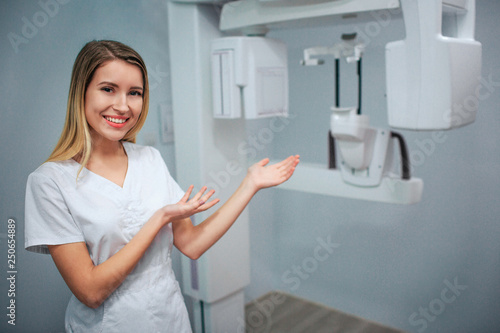 Happy positive young dentist stand in x-ray room and point on machine. Alone in white robe.
