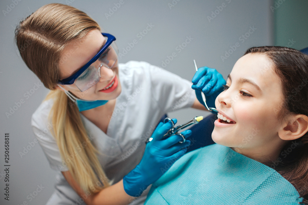 Careful female dentist hold tools close to girl mouth. Child show front teeth. She sit calmly in dental chair. Careful female dentist hold tools close to girl mouth. Child show front teeth. She sit calmly in dental chair.