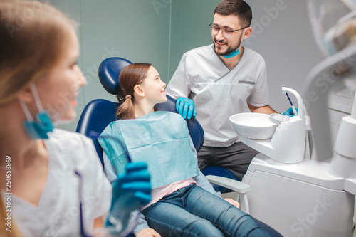 Child sit in denta lchair in room. Male dentist stand beside her and smile. Young woman look at them. She smile too.