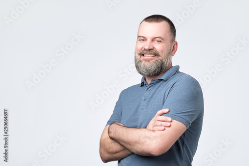 Fotografia Mature man with beard holding hands crossed and looking at camera while smiling