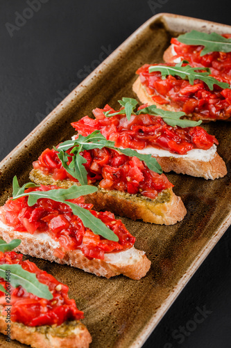Canvas Print Appetizer bruschetta with chopped vegetables on ciabatta bread and sauce pesto on plate over black stone background
