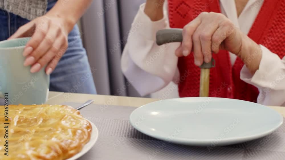 Tracking close-up shot of unrecognizable senior woman with stick sitting at table while granddaughter pouring tea into mug for her; there is delicious pie and buns on table Tracking close-up shot of unrecognizable senior woman with stick sitting at table while granddaughter pouring tea into mug for her; there is delicious pie and buns on table