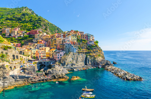 Manarola traditional typical Italian village in National park Cinque Terre, colorful multicolored buildings houses on rock cliff, fishing boats on water, blue sky background, La Spezia, Liguria, Italy