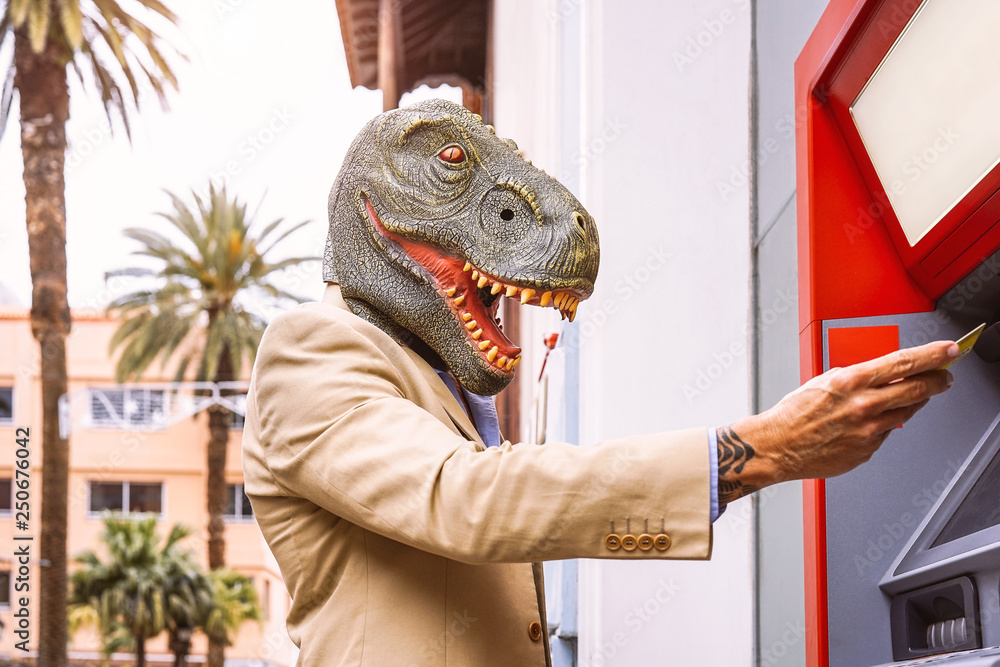 Senior man wearing t-rex dinosaur mask withdraw money from bank cash machine with debit card - Surreal image of half human and animal - Absurd and crazy concept of ATM advertise