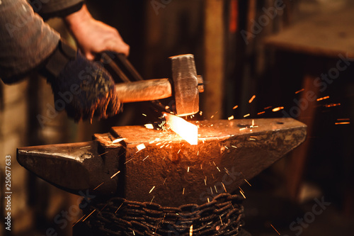 Fotografie, Obraz The blacksmith manually forging the red-hot metal on the anvil in smithy with spark fireworks