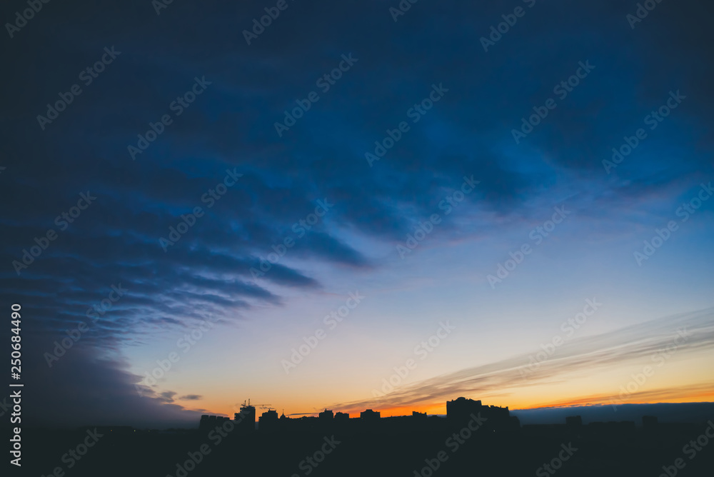 Fototapeta premium Cityscape with wonderful varicolored vivid dawn. Amazing dramatic blue cloud sky above dark silhouettes of city building roofs. Atmospheric background of orange sunrise in overcast weather. Copy space