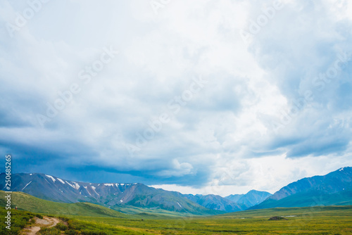 Spectacular view of distant giant mountains. Footpath through valley in highlands. Hiking path. Wonderful huge mountain range under cloudy sky. Amazing dramatic green landscape of majestic nature.