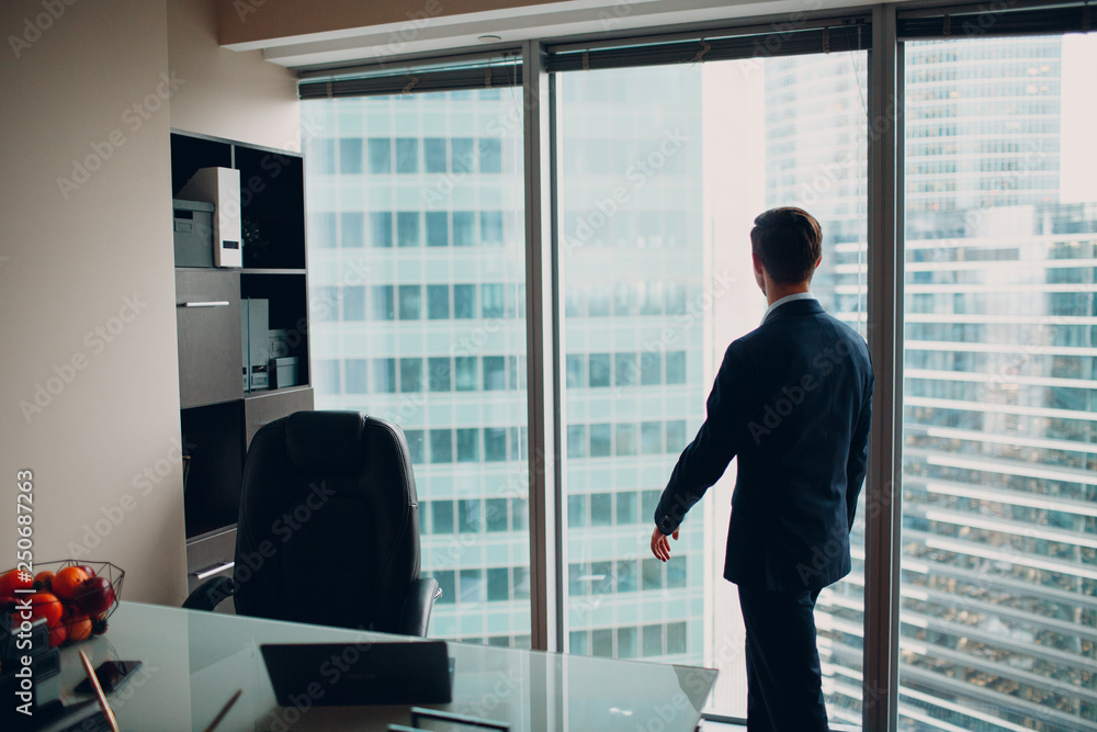 Fototapeta premium A young man in a skyscraper office is standing by a panoramic window.