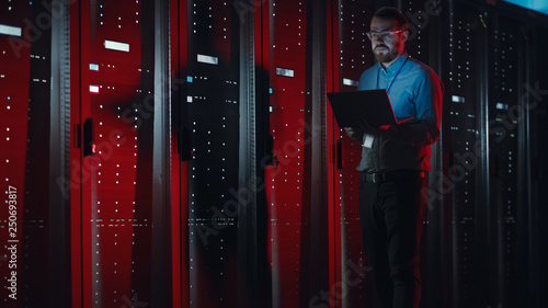 Fotografie Bearded IT Specialist in Glasses is Working on Laptop in Data Center while Standing Near Server Rack
