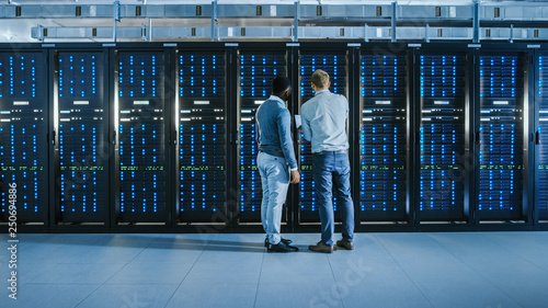 Obraz na plátně Bearded IT Technician in Glasses with a Laptop Computer and Black Male Engineer Colleague are Talking in Data Center while Working Next to Server Racks