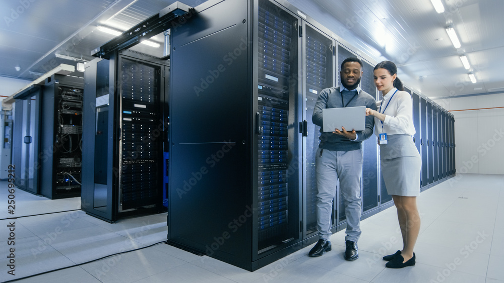 Black IT Technician with a Laptop Computer Gives a Tour to a Young Intern. They Talk in Data Center while Walking Next to Server Racks. Running Diagnostics or Doing Maintenance Work. Black IT Technician with a Laptop Computer Gives a Tour to a Young Intern. They Talk in Data Center while Walking Next to Server Racks. Running Diagnostics or Doing Maintenance Work.