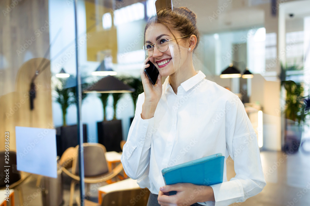 Stylish young businesswoman talking with her mobile phone while holding her digital tablet standing in the hotel lobby.