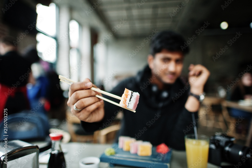 Close up photo of sticks holding casual and stylish young asian man with earphones at cafe eating sushi.
