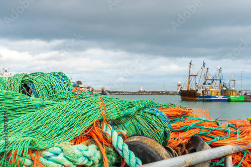 Stacked green and orange coloured fishing nets with fishing boats in the background docked at Howth Harbour in County Dublin, Ireland.