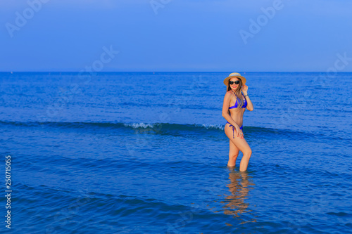 girl in a blue swimsuit, sunglasses and boater poses in seawater