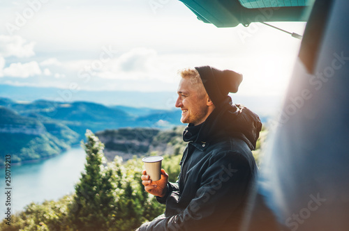 hipster tourist hold in hands mug of hot drink, lonely guy smile enjoy sun flare mountain in auto, happy traveler drink cup of tea on nature, vacation weekend concept on panoramic landscape