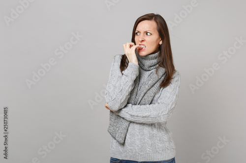 Preoccupied young woman in gray sweater, scarf looking aside, gnawing nails isolated on grey wall background. Healthy fashion lifestyle people sincere emotions cold season concept. Mock up copy space.