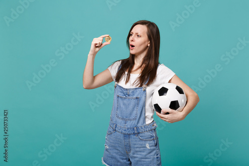 Perplexed young woman football fan support favorite team with soccer ball, bitcoin future currency isolated on blue turquoise wall background. People emotions, sport family leisure lifestyle concept. © ViDi Studio